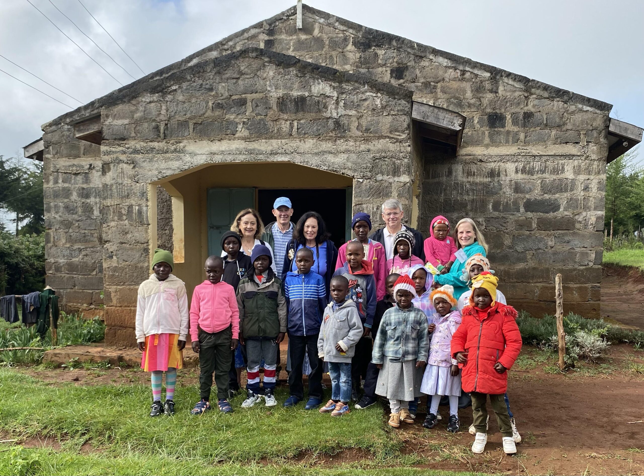 Group of children and adults in front of a stone building in Kenya, showcasing the community involvement of Lakewood Methodist Church's mission initiative.
